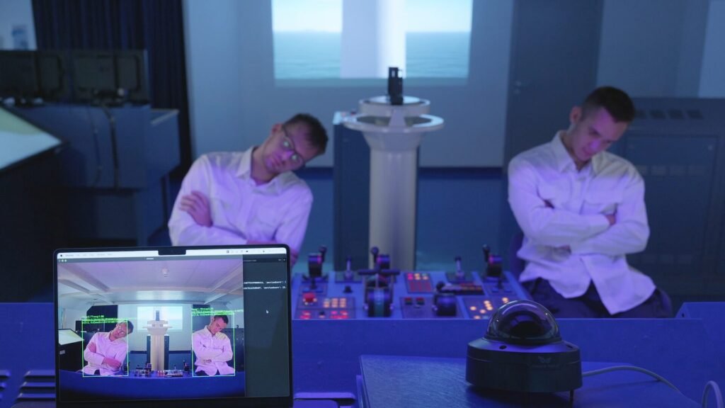 Two men in white shirts sit with folded arms, heads drooped in sleep at a blue-lit ship-bridge simulator; a laptop in the foreground shows surveillance software highlighting their slumped posture, with control panels and a simulated ocean view in the background.