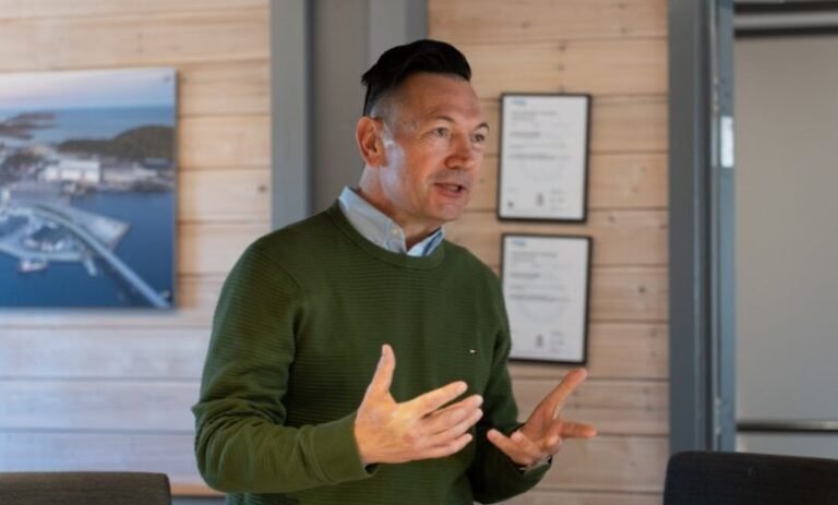 Man in a green sweater speaks and uses hand gestures during a meeting in a wood-paneled room.